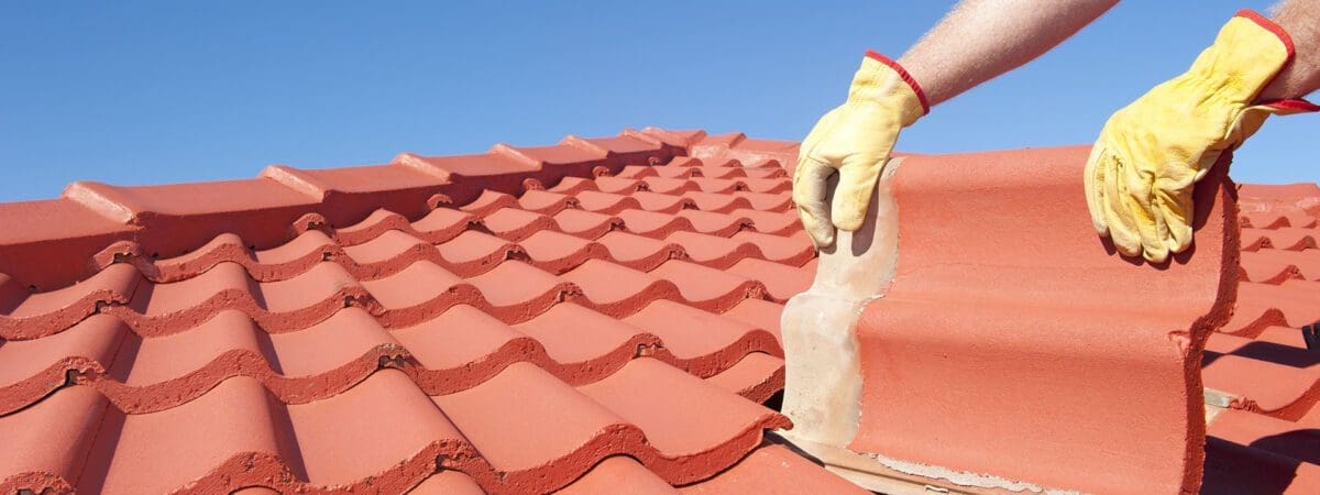 Roof repair worker with yellow gloves replacing red tiles or shingles on house with blue sky as background and copy space.